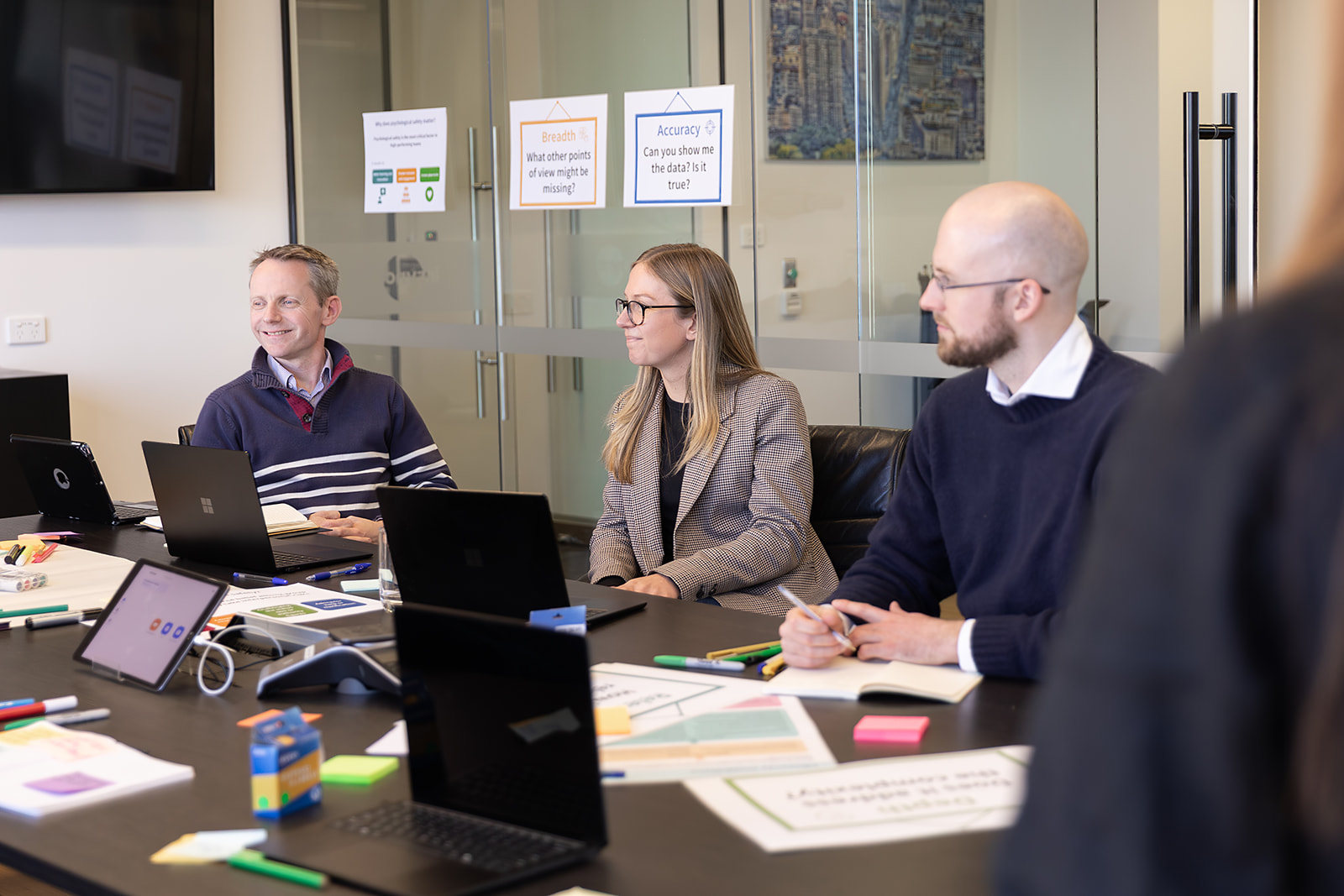 Three professionals sit around a conference table during a meeting, with laptops, papers, and markers on the table and glass walls with posters behind them.