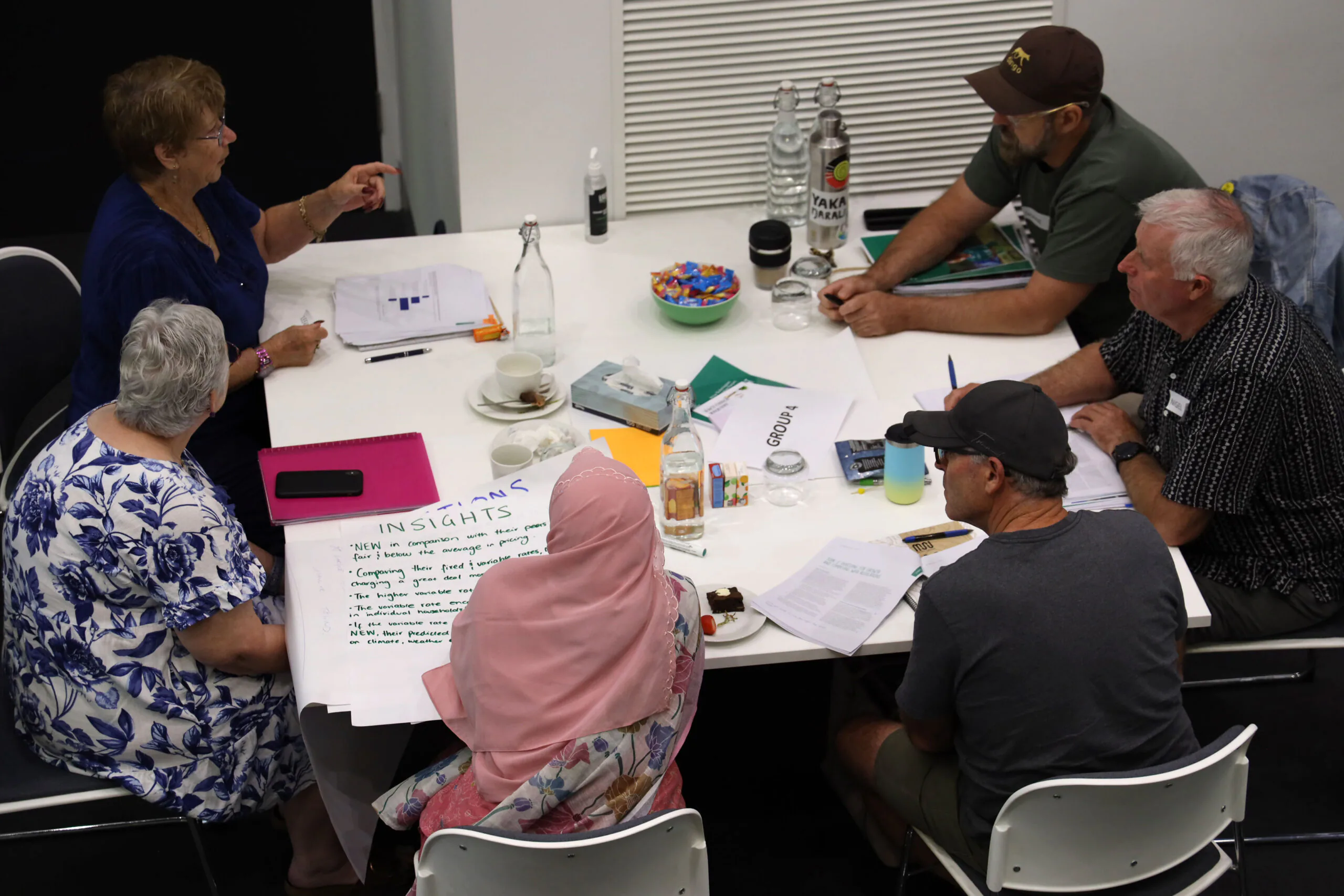 Group of adults around a white table in a discussion, with papers, pens, cups, water bottles and snacks visible on the table.